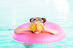 快乐的孩子在游泳池里用充气环游泳时, 眼睛附近拿着半橘子