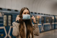 一名妇女戴上医疗面罩，以避免在地铁站传播高头牛病毒。一个穿着长发外套的女孩正在地铁站的月台上等火车.