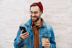 一个英俊、快乐、时髦、留着胡子的年轻人走在街上，手里拿着一杯外卖咖啡，用的是手机