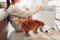 冬天在家使用加热器。妇女温暖她的手坐在设备与猫和穿着温暖的衣服。加热季节.