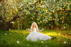 日落时分, 一个穿着白色衣服的小女孩站在苹果树下, 面带微笑。花瓣在飞翔