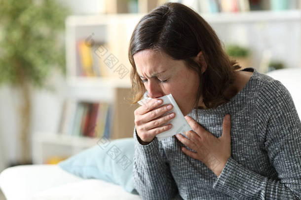 生病的成年妇女坐在家里的沙发上，<strong>用纸</strong>巾捂住嘴咳嗽