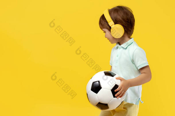 在黄色背景下，可爱的小男孩拿着<strong>足球</strong>，用耳机听音乐的特写照片.