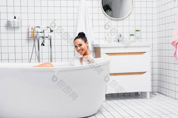 快乐的黑发<strong>女人</strong>一边用泡沫洗澡一边看着相机 