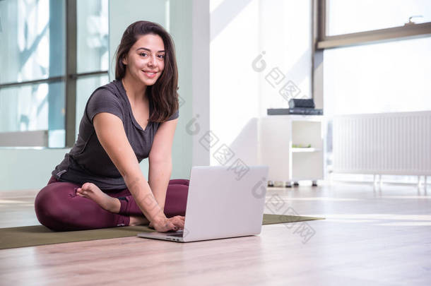 在潘德米亚期间，年轻女子用笔记本电脑练习<strong>瑜伽</strong>的照片。漂亮的女孩在<strong>课</strong>堂上练习<strong>瑜伽</strong>。<strong>瑜伽</strong>工作室指导员。模糊的背景