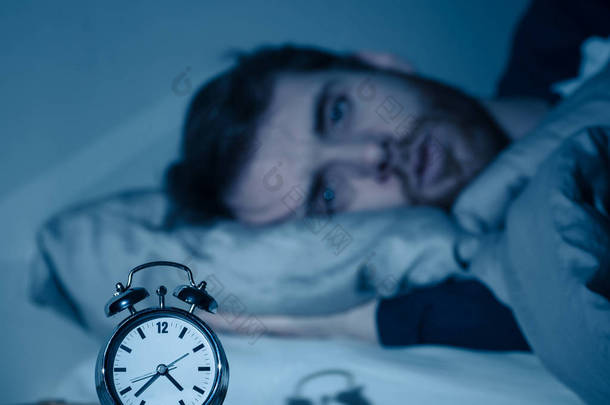 失眠和绝望的<strong>年轻</strong>白种人男子晚上醒来不能睡觉,感到沮丧和担心看着时钟遭受失眠的压力和睡眠障碍的概念.
