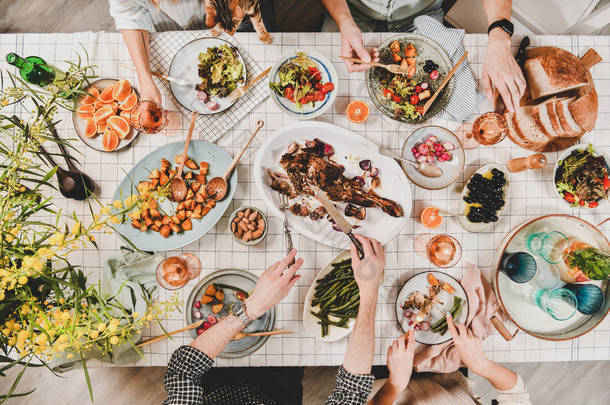 家人或朋友聚餐。平装人的手与烤羊肩, 沙拉, 蔬菜, 葡萄酒, 含羞草树枝在白色格子桌布, 顶视图。庆祝派对晚宴
