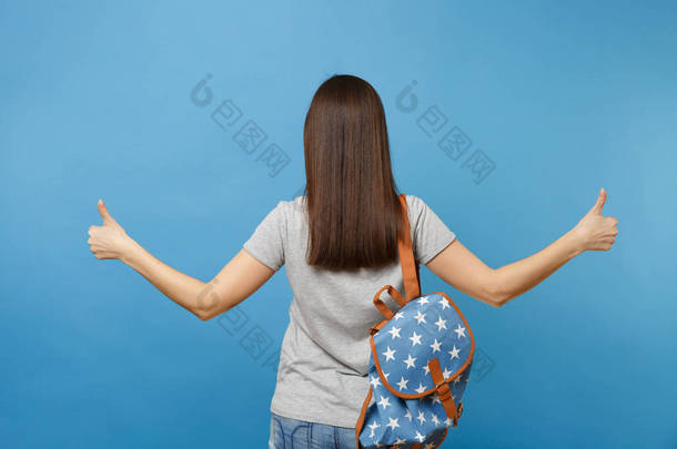 <strong>背部</strong>后视图的年轻黑发女子的学生在休闲服装与背包站立显示竖起大拇指在蓝色背景上孤立。大学教育。复制广告空间