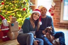 年轻漂亮的夫妇面带微笑，既快乐又自信。 在家里，坐在地板上，头戴圣爪帽，抱着狗绕着圣诞树