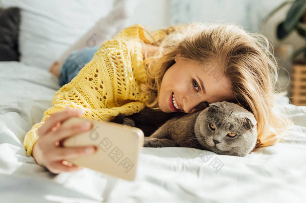 美丽的微笑的年轻女子在智能手机上自拍, 而躺在床上与苏格兰折叠猫 
