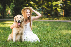 美丽的年轻女孩在白色礼服触摸草帽，同时拥抱金毛猎犬，而微笑，坐在草地上，看着远方