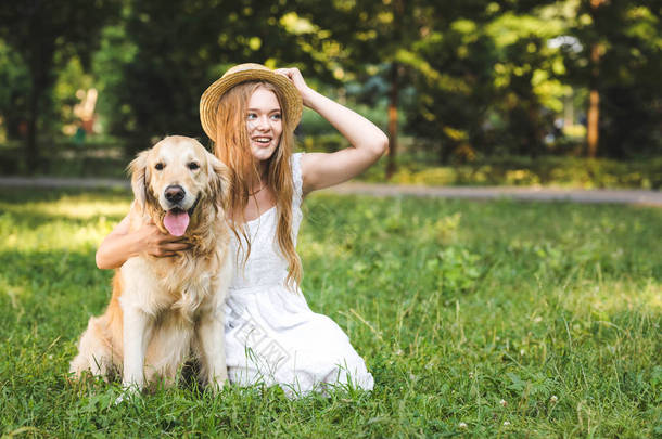 美丽的年轻女孩在白色礼服触摸草帽，<strong>同时</strong>拥抱金毛猎犬，而微笑，坐在草地上，看着远方