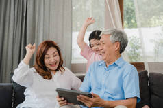 亚洲祖父母和孙女在家里用平板电脑.上了年纪的中国人，爷爷奶奶高兴地和小女孩一起在社交媒体上消磨时光，躺在沙发上享受客厅的概念