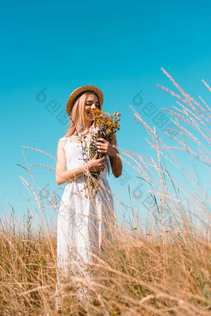 金发碧眼的女人，戴着草帽，身穿白衣，手持野花束迎着蓝天