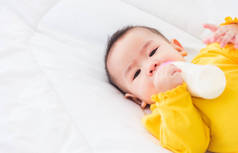 亚洲漂亮的小女孩穿着一件黄色的衣服，吃着睡在白色床上的牛奶，抱着一瓶牛奶的婴儿，婴儿食品的概念