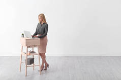 使用膝上型计算机的年轻妇女站在工作场所反对白墙。文本空间