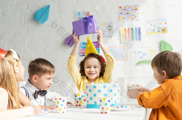 快乐的孩子拿着礼物, 而坐在桌子上与朋友在生日聚会庆祝活动