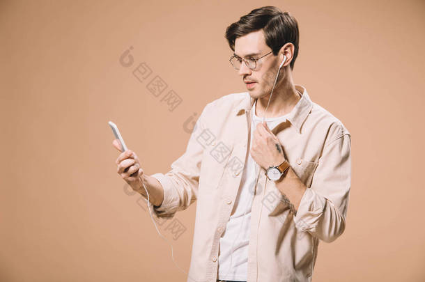 英俊的男人在眼镜看智能手机, 而听音乐在耳机隔离米色 