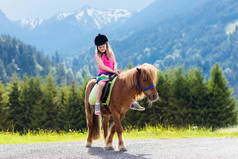 孩子们骑着小马在阿尔卑斯山里在奥地利蒂罗尔的马场度过家庭春假。孩子们骑马。孩子照顾动物。孩子和宠物。在盛开的草地上的小女孩.