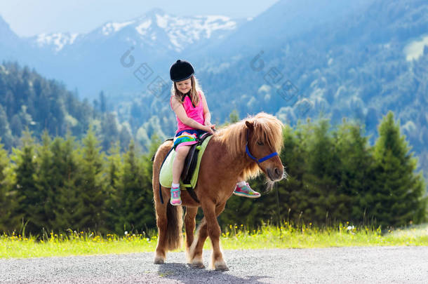 孩子们骑着小马在阿尔卑斯山里在奥地利蒂罗尔的马场度过家庭春假。孩子们骑马。孩子照顾动物。孩子和宠物。在盛开的<strong>草地</strong>上的小女孩.