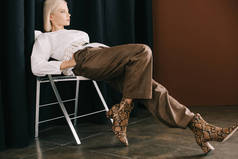时尚金发女人在白色上衣和靴子与蛇皮打印坐在椅子上接近棕色的窗帘