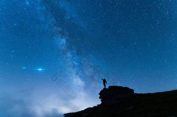 <strong>一</strong>个站着的年轻人在山上高举双臂的轮廓。带着蓝夜星空旅行的背景.