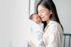 白衬衫亚洲母亲亲吻她的新生儿在卧室前的玻璃窗与白色窗帘，以显示爱和家庭纽带.