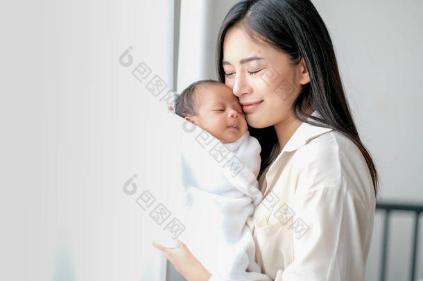 白衬衫亚洲母<strong>亲亲</strong>吻她的新生儿在卧室前的玻璃窗与白色窗帘，以显示爱和家庭纽带.