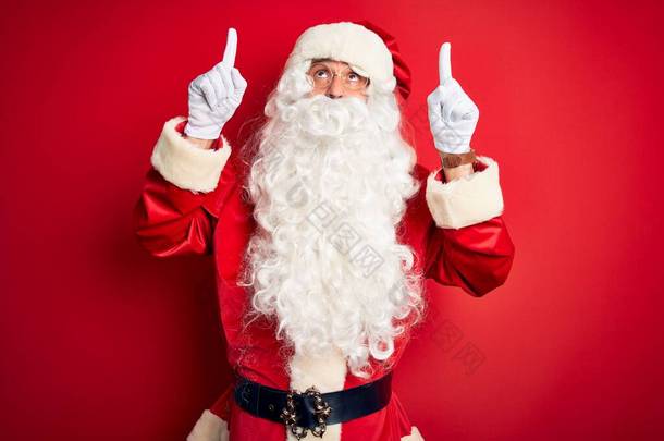 中年男子穿着圣诞老人的服装，站在孤立的红色背景上，面带微笑，惊讶，用手指和胳膊指指着.
