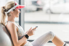 愉快的女运动员与智能手机的侧视图使用智能手机附近的健身球在健身房