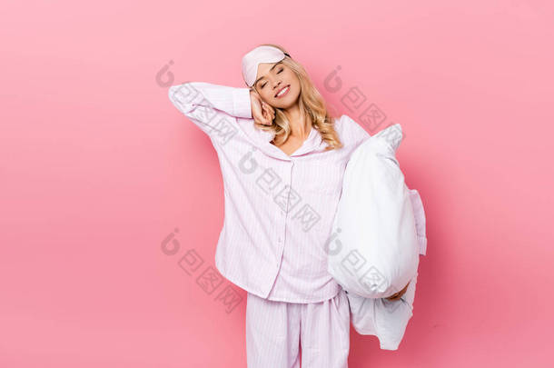 穿着睡衣、蒙着<strong>眼睛</strong>、拿着枕头、伸展着粉红背景的女人微笑