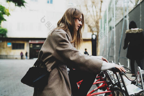 时尚的学生女孩穿着时髦的衣服坐在租用的城市自行车, 并准备骑上 