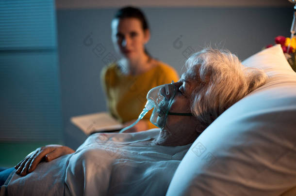 医院的一位女士在晚上帮助一位戴氧气面罩的老年病人，她正在看书，检查她的<strong>病情</strong>