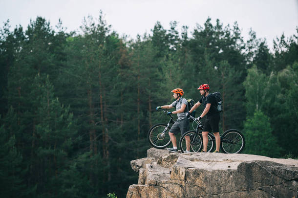 年轻的试验骑自行车站在岩石悬崖上的侧面看着模糊的松树林背景和指向某处