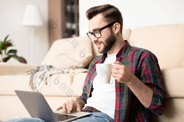 年轻的散工，戴着茶杯和笔记本电脑，坐在客厅的沙发上，孤身一人工作