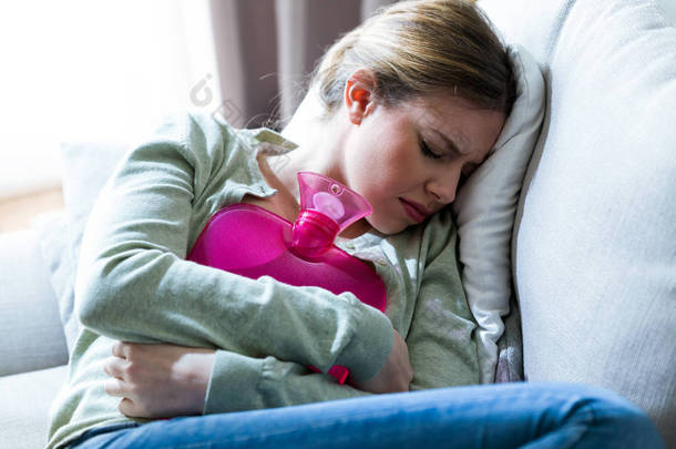 不健康<strong>的</strong>年轻妇女胃痛使用热水袋, 而坐在沙发上在家.