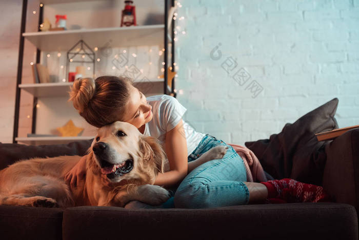 年轻的金发碧眼的女人在沙发上拥抱金毛猎犬狗在圣诞节时间