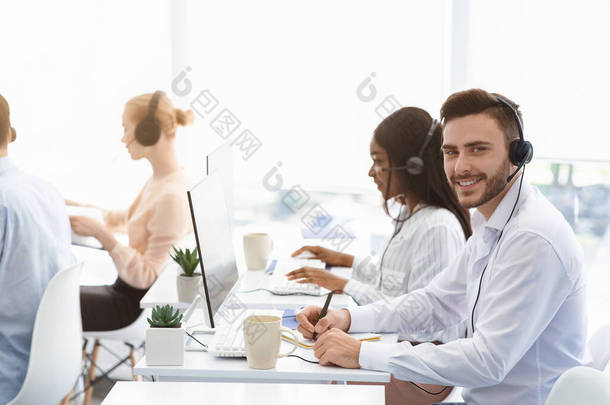 吸引人的呼叫<strong>中心</strong>操作员对着摄像机微笑，他的同事在办公室和客户交谈，复制空间