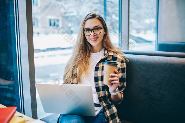 现代技术和通信的概念, 成功的女文案员坐在室内, 手里拿着一杯外卖咖啡, 腿上拿着现代笔记本电脑看着相机