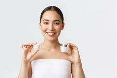 美容美发、个人护理、温泉沙龙及护肤理念.漂亮的亚洲女人穿着浴巾，脸上有两种乳霜和眼霜，笑着推荐使用水合作用，滋养肌肤护理疗法