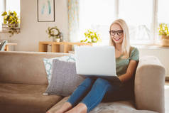 微笑的年轻金发碧眼的女人坐在家里的沙发上, 使用笔记本电脑.