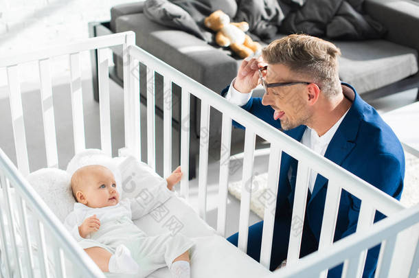 幸福的父亲在正式的服装显示舌头对幼女在婴儿床 