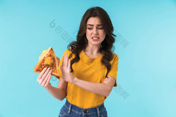 照片上年轻美丽的女子举着三明治站在蓝色墙壁上，显得很不高兴，她举手投足.