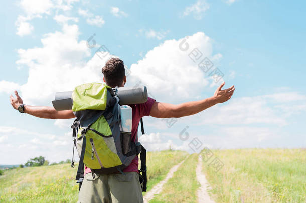 徒步旅行者与背包和旅游垫站立在绿色草甸伸出的手的后面看法