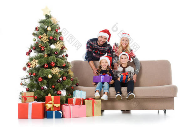 微笑的家庭与孩子在圣诞老人的帽子坐在沙发附近的圣诞树与礼品盒, 孤立的白色