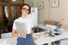 美丽的女孩在眼镜和白色 t恤朦胧地看着相机手持手提电脑在现代办公室工作