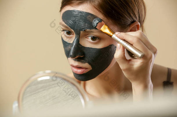 焦炭面罩或黑色粘土泥. 美丽的女人正在她的脸上涂上洁白的黑色面具,近距离拍摄工作室镜头. 家庭温泉天然化妆品的概念.