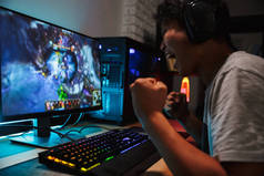 快乐的亚洲青少年玩家玩电子游戏的电脑在黑暗的房间戴着耳机, 并使用背光彩色键盘