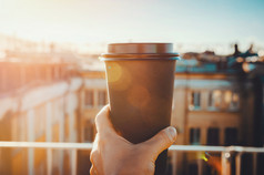手拿着热工艺杯咖啡或茶在早晨的阳光，看模糊的城市背景。享受，生活方式，带走早餐的概念。女人在屋顶上喝酒