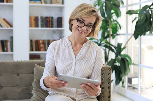 快乐的中年妇女坐在沙发上，使用电脑平板电脑应用程序，观看屏幕，在社交网络上阅读好消息，在网上购物或聊天